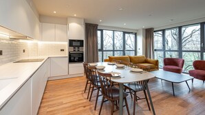 A modern kitchen with white cabinets, a dining area with a round table and chairs, and a large window with a view of trees. - Spacious Flat in North London (London)
