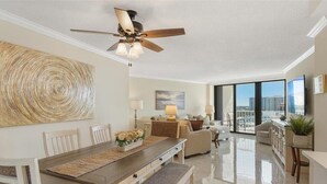 A spacious living area with a dining table, chairs, a sofa, and a large window offering a city view. - Beachfront 2BR/2BA condo with pool, WiFi, and Marco Island views. (Marco Island)