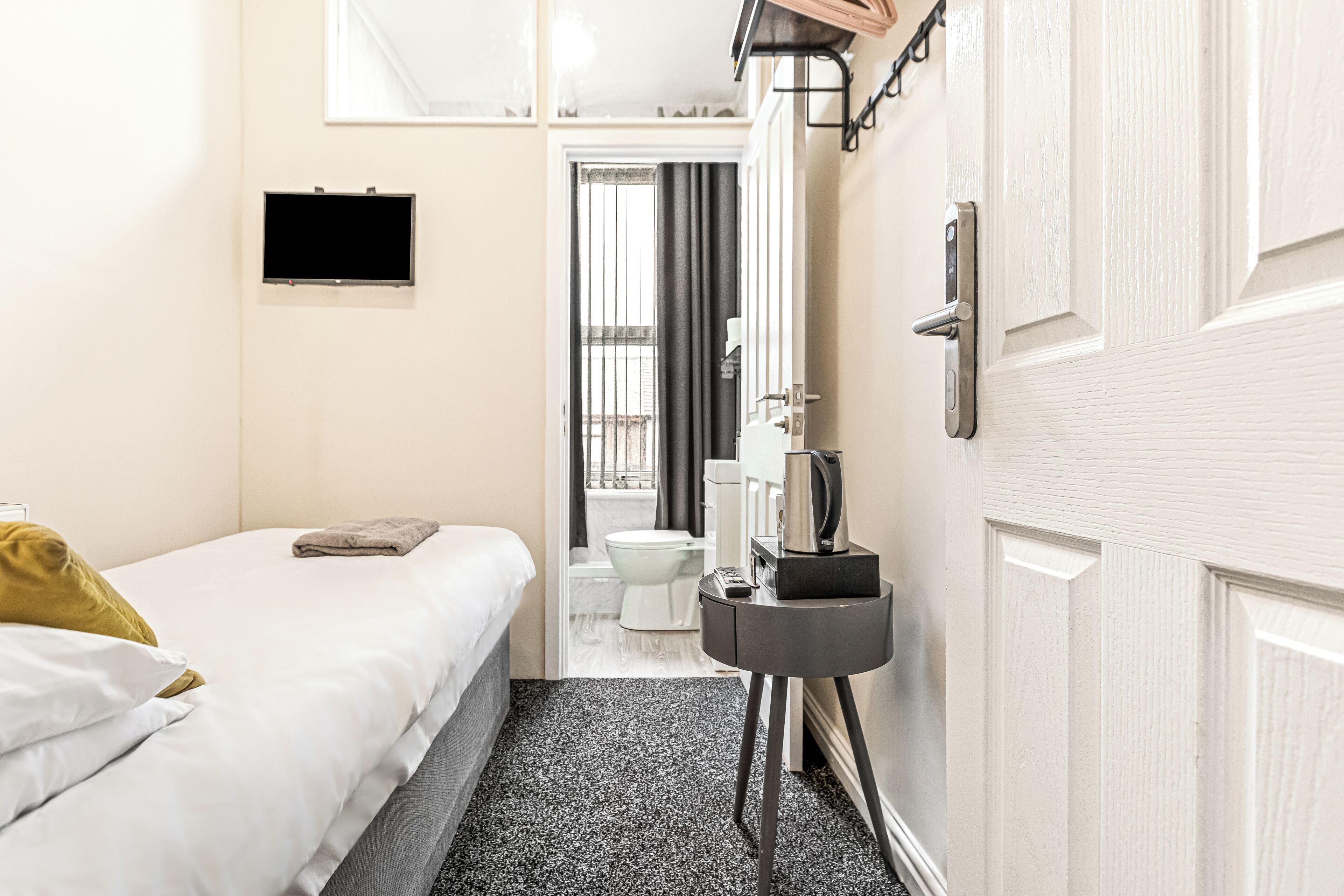 A hotel room with a bed, a small table with a kettle, a TV, and a door leading to a bathroom.