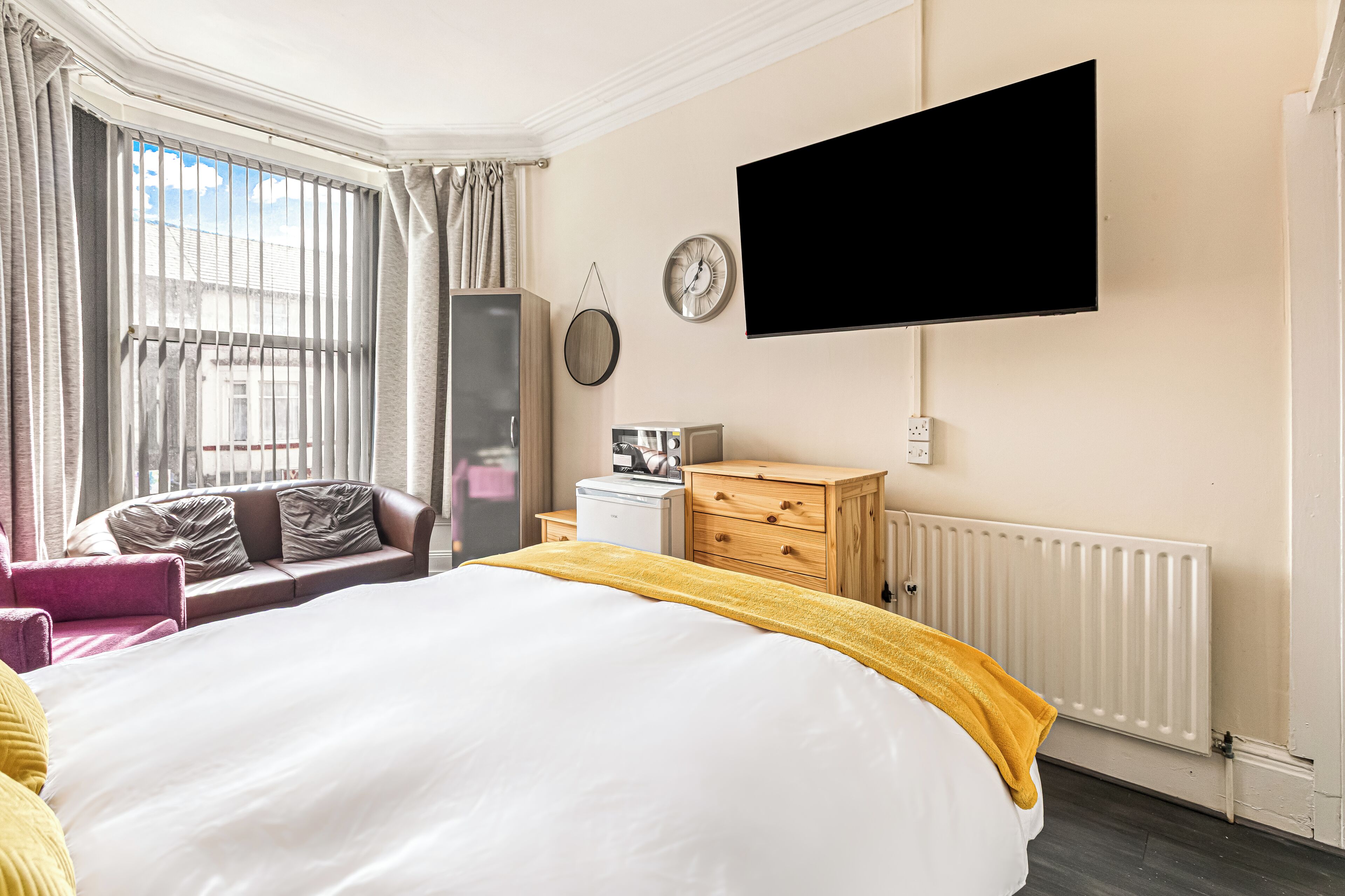 A hotel room with a bed, a TV mounted on the wall, a dresser, a nightstand, a window with curtains, and a radiator.