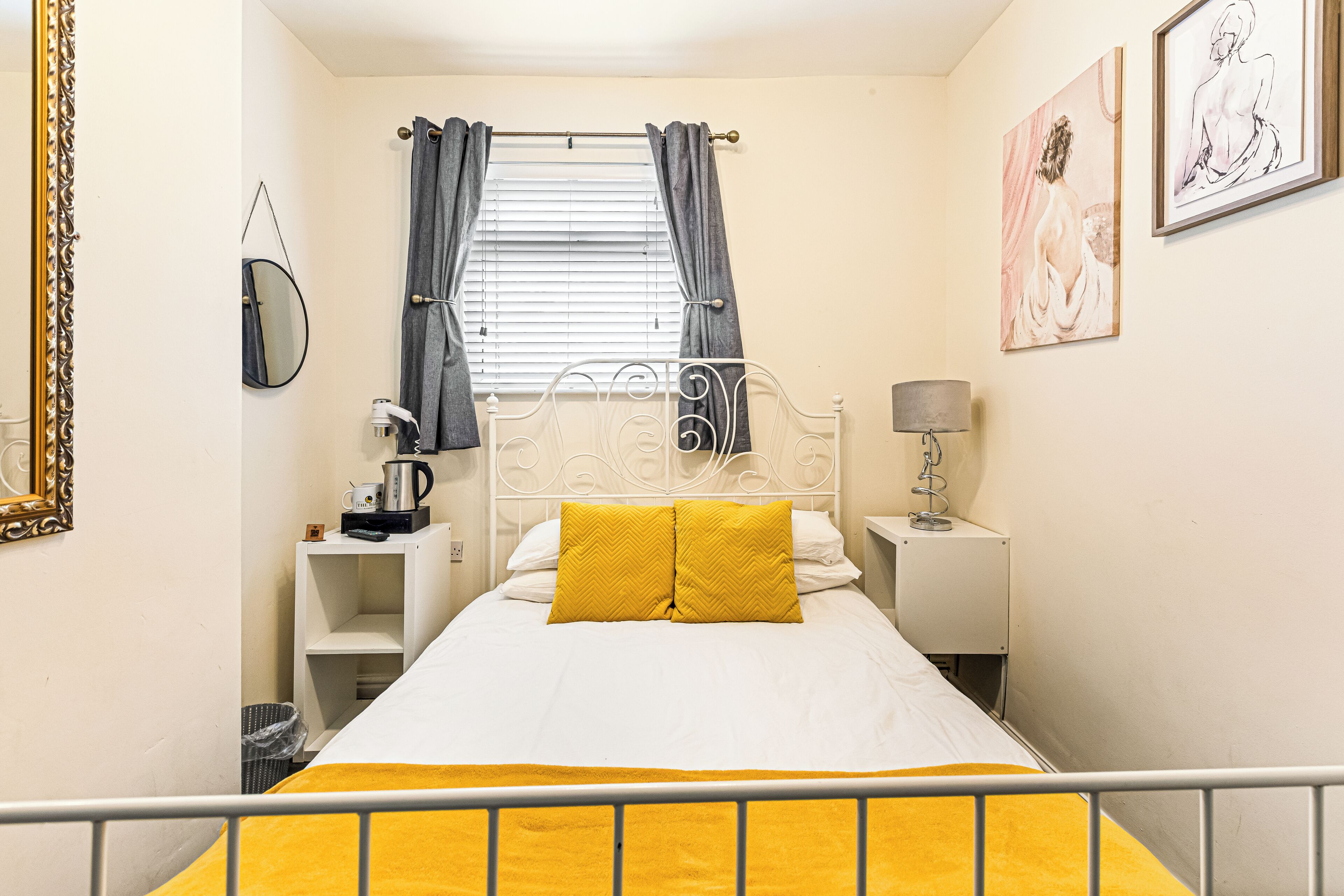 A bedroom with a bed, a bedside table, a lamp, a window with curtains, a mirror, and artwork on the wall.