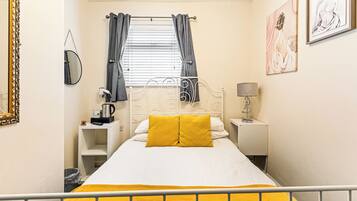 A bedroom with a bed, a bedside table, a lamp, a window with curtains, a mirror, and artwork on the wall.