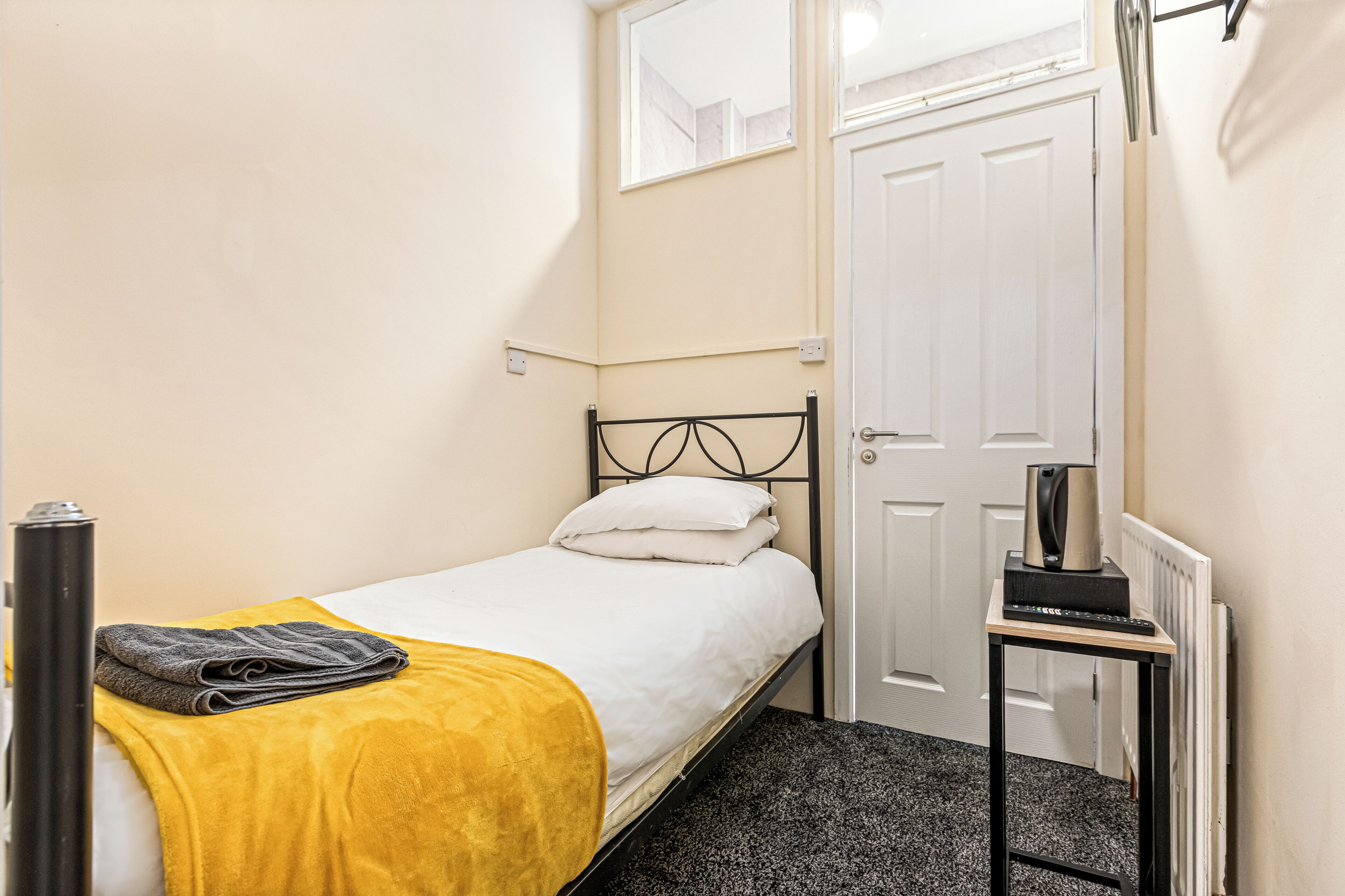 Ein kleines Zimmer mit einem Einzelbett, einer weißen Tür, einem kleinen Tisch mit Wasserkocher und einem Fenster über dem Bett.