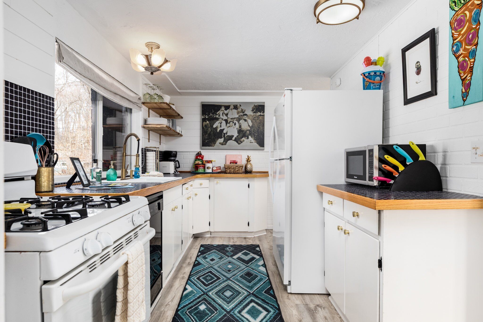 A compact kitchen with white cabinets, a gas stove, a microwave, and a sink. There is a patterned rug on the floor and a painting on the wall.