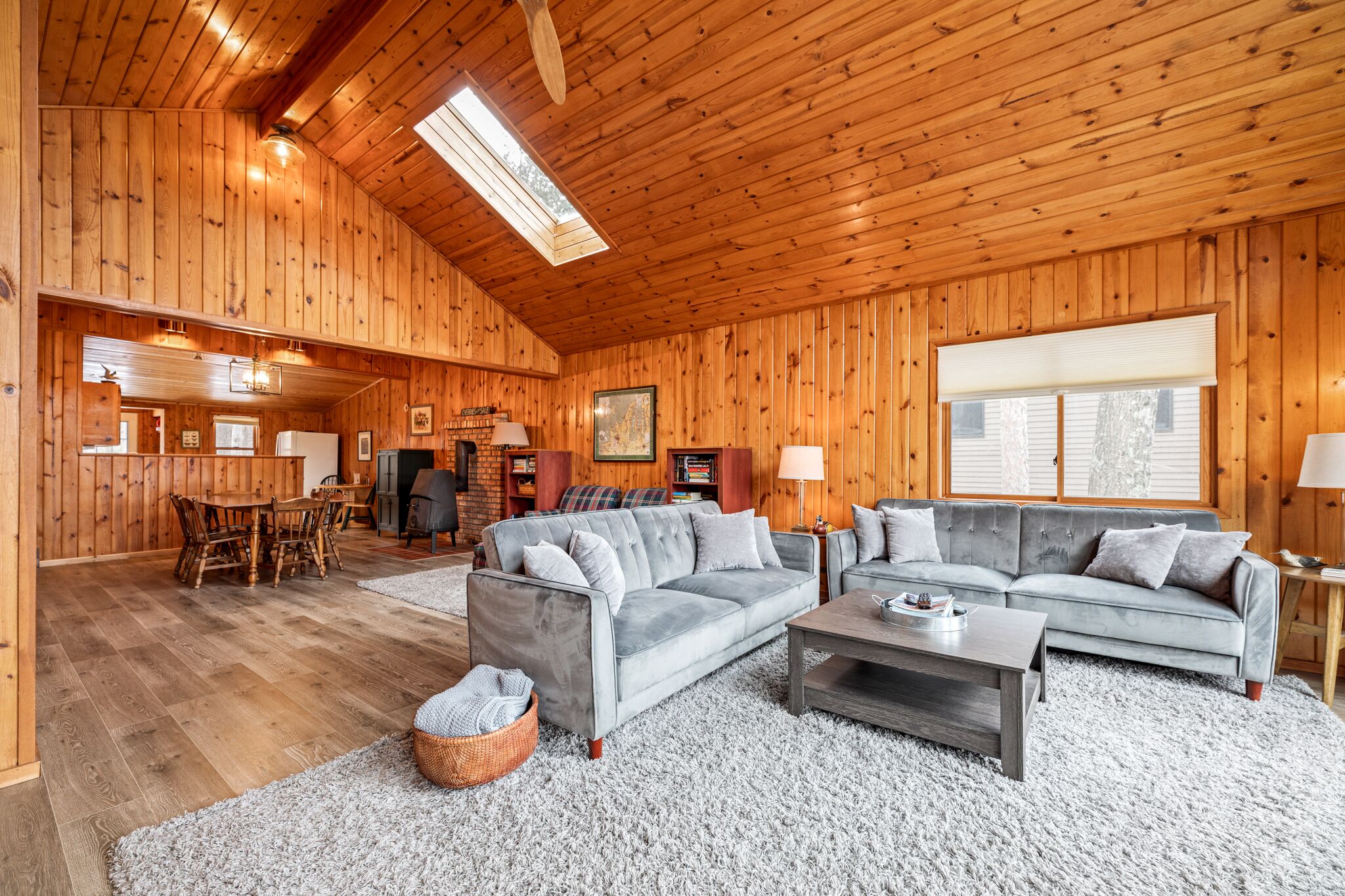 A spacious living room with wooden walls and a high ceiling, featuring a grey sofa, a coffee table, and a dining area with a wooden table and chairs.