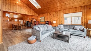 En rummelig stue med trævægge og højt til loftet, med en grå sofa, et sofabord og et spiseområde med et træbord og stole.