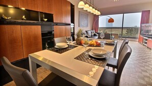 A modern dining area with a long table, chairs, and a kitchen in the background.