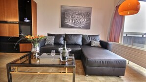A modern living room with a glass coffee table, a black leather sofa, and a large window with curtains.