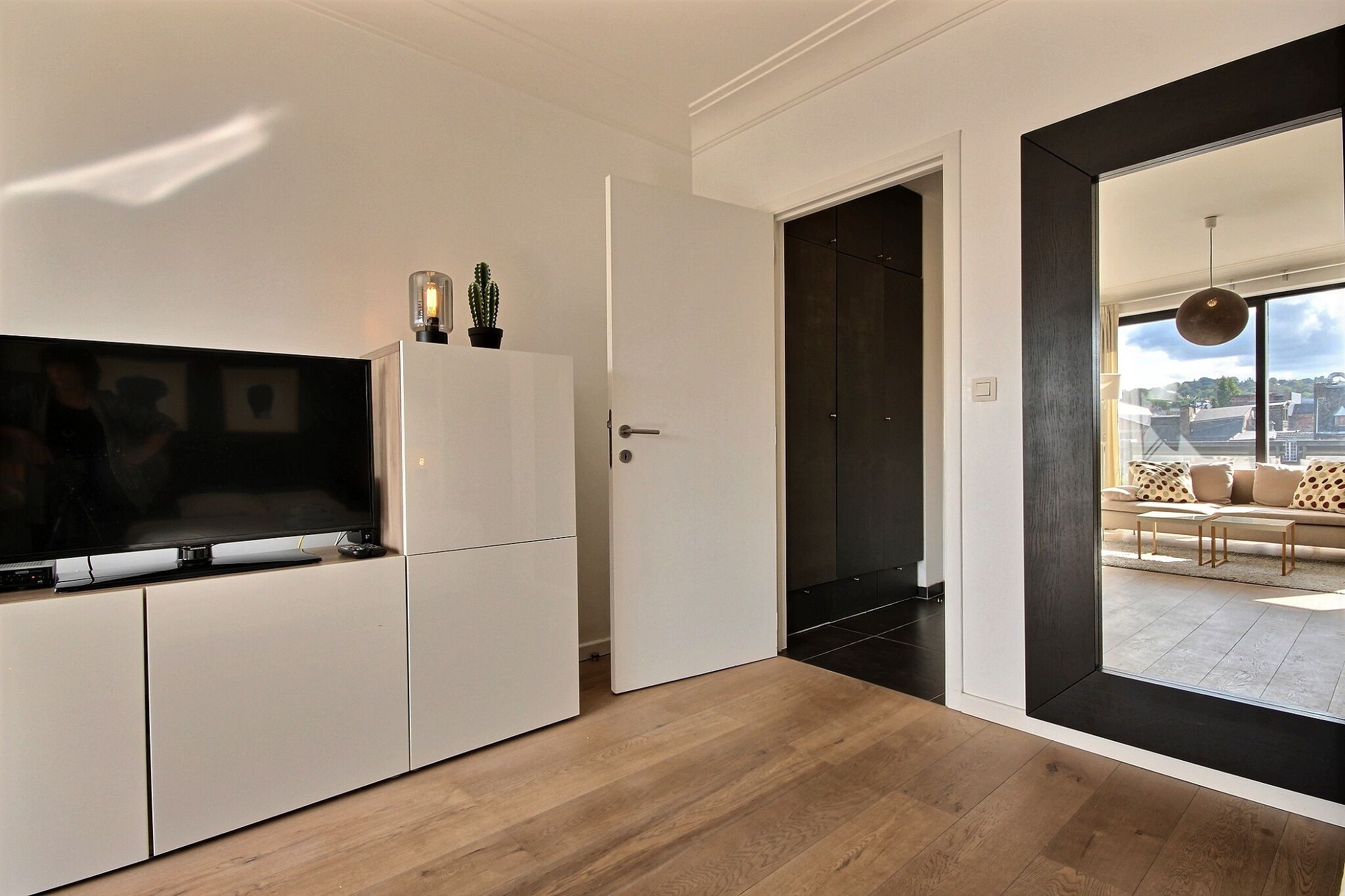 A modern living room with a flat-screen TV, a door leading to a balcony, and a view of the cityscape.