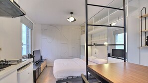Moderni makuuhuone, jossa on sänky, yöpöytä ja seinään upotettu televisio. Huoneessa on myös keittiöalue, jossa on liesi ja jääkaappi. Huoneessa on suuri ikkuna ja peili seinällä.