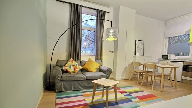 A modern living room with a sofa, a small table, a colorful rug, and a dining area with chairs and a table. - Li Tore 1 - Nice practical studio in the center (Liège)