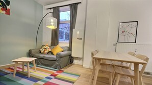 A modern living room with a grey sofa, a colorful striped rug, a wooden dining table, and a large window with curtains. - Li Tore 1 - Nice practical studio in the center (Liège)