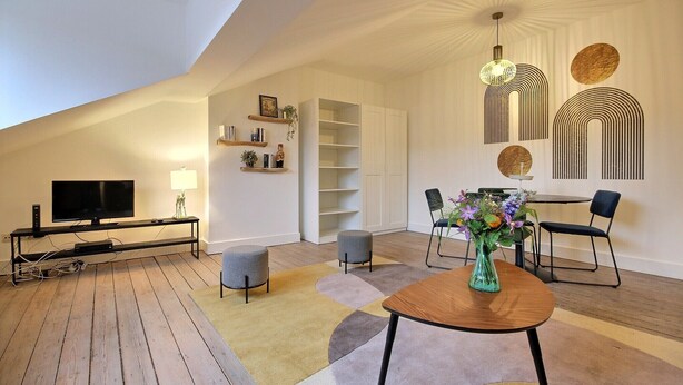A modern living room with a TV, sofa, ottomans, a dining table, and a vase of flowers.