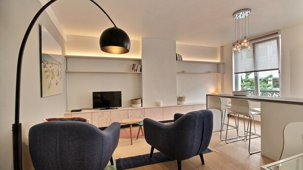 Ein modernes Wohnzimmer mit Fernseher, zwei Sofas, einem Essbereich mit Tisch und Stühlen sowie einer Küchenzeile mit Fenster.
