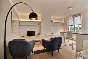 Een moderne woonkamer met een televisie, twee banken, een eethoek met tafel en stoelen, en een keuken met een raam.