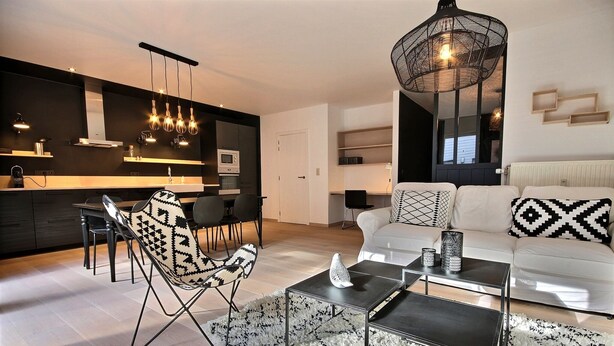 A modern living room with a sofa, coffee table, and patterned chairs. A kitchen with a dining area is visible in the background. - Chez Rémy - 3 bedrooms (Liège)