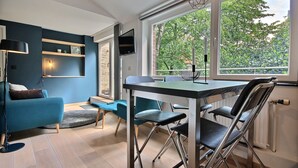 A modern living room with a blue sofa, a dining table with chairs, a TV mounted on the wall, and a large window with a view of trees. - Li Tore 3 - Pretty modern studio in the center (Liège)