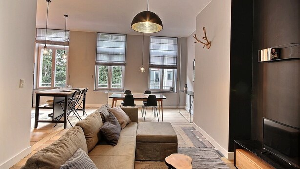 Een moderne woonkamer met een bank, eettafel, stoelen, grote ramen en een flatscreen televisie.