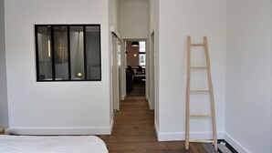 Een smalle gang met een houten vloer, een raam met zwarte kaders, en een houten ladder die tegen de muur staat.