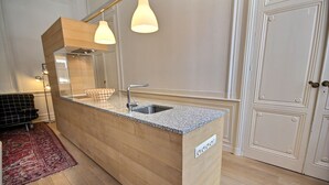 A modern kitchen with a granite countertop, stainless steel sink, and wooden cabinetry. There is a large mirror above the sink and pendant lighting.