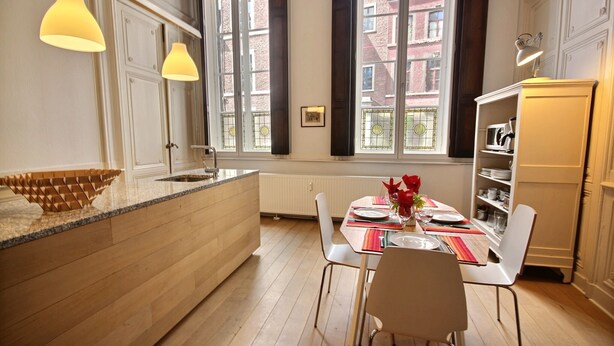 A modern kitchen with a central island, pendant lighting, and a view of a red brick building through large windows.