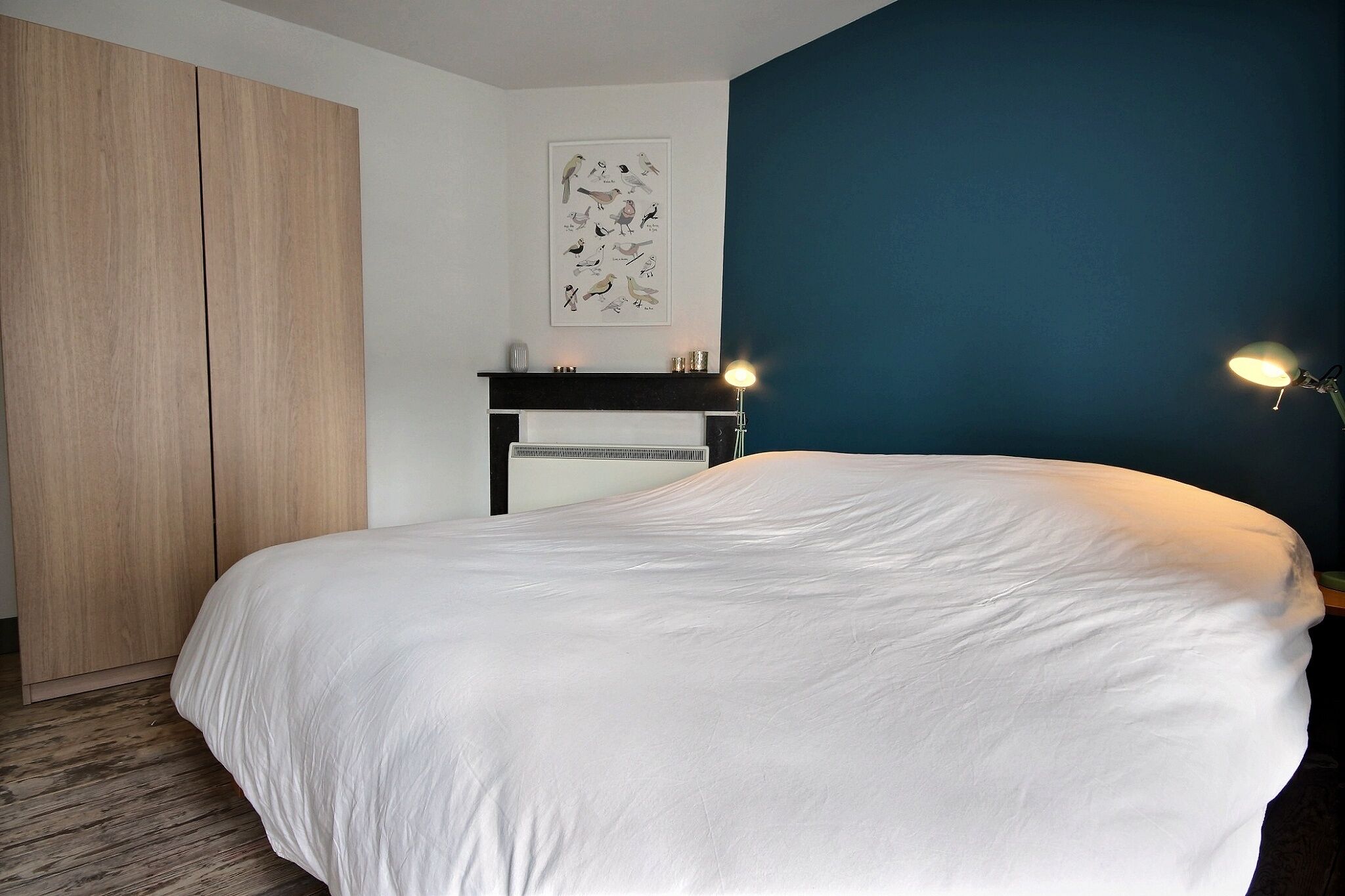 A bedroom with a large bed, a wooden wardrobe, a fireplace, and a wall-mounted artwork.