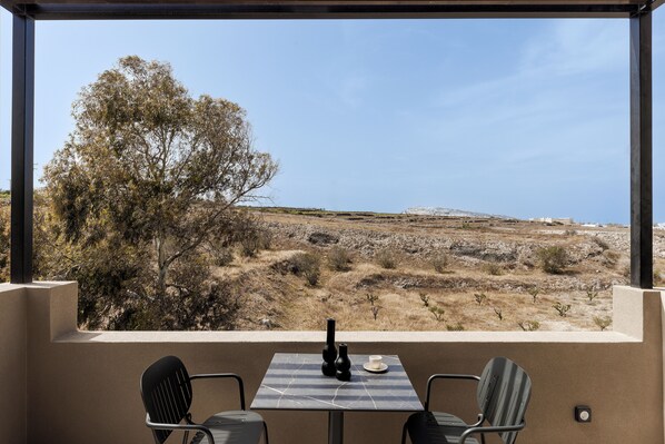 Un balcon avec une table et des chaises, donnant sur un paysage rocheux à la végétation clairsemée et offrant une vue lointaine sur un bâtiment.
