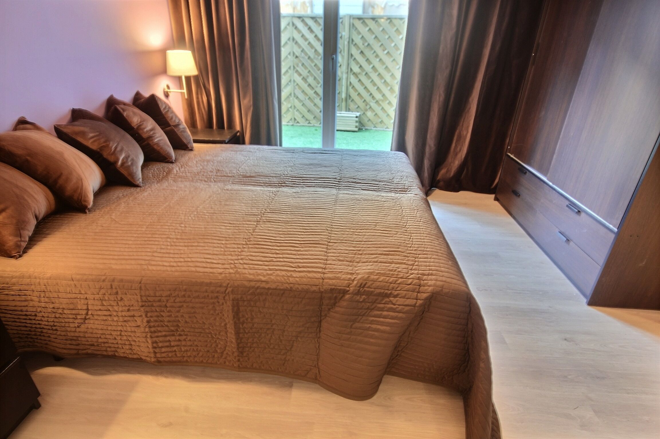 Une chambre avec un grand lit, une table de chevet avec une lampe et une vue sur un espace extérieur clos.