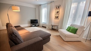 Una sala de estar moderna con un sofá seccional marrón, un sofá blanco con almohadas verdes, un televisor de pantalla plana y una lámpara de pie.