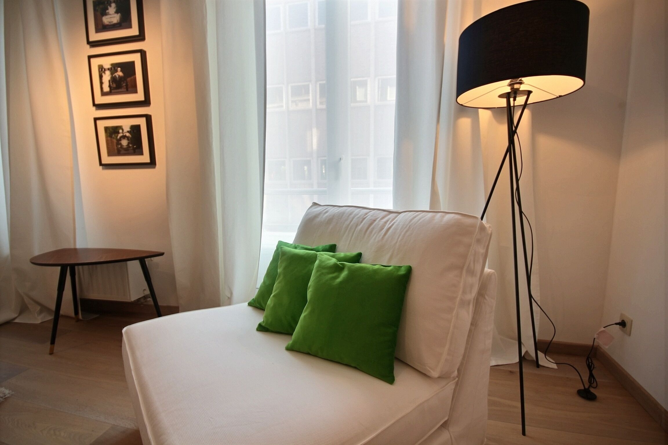 Habitación de hotel con cama, almohadas verdes, una lámpara de pie, una mesita pequeña y cuadros en la pared.