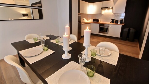 Un comedor con una mesa de madera oscura dispuesta para cuatro personas, platos blancos, vasos verdes y velas blancas. Se ve una cocina al fondo.