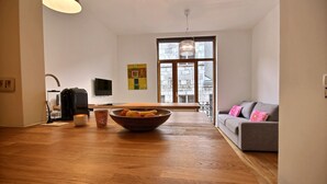 A modern living room with a wooden floor, a grey sofa, a large window, and a bowl of fruit on a table. - Goffe Street - Studio (Liège)
