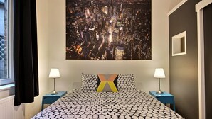 Une chambre d’hôtel avec un grand lit, deux tables de chevet avec des lampes, une œuvre d’art représentant un paysage urbain accrochée au mur, et une fenêtre avec des rideaux.