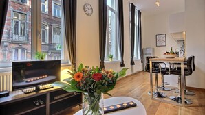 Un salon moderne doté d’un téléviseur à écran plat, d’une table à manger avec des tabourets de bar, d’un vase rempli de fleurs et de grandes fenêtres avec des rideaux.