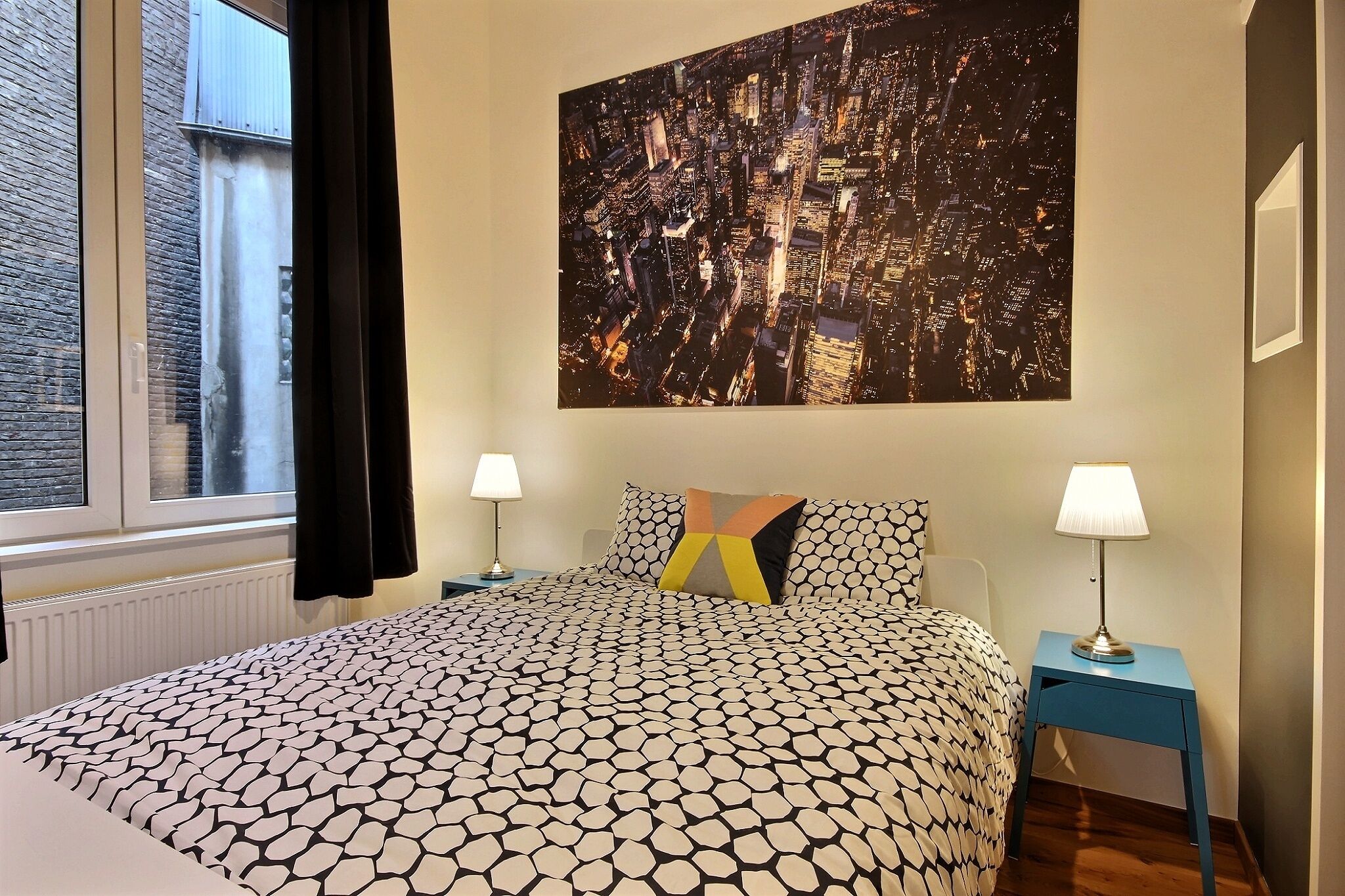 Ein ordentlich gemachtes Bett mit einem gemusterten Bettüberwurf, zwei Nachttischlampen und einem großen Stadtbild an der Wand.