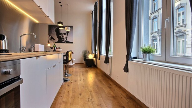 Une cuisine étroite avec un plancher en bois, un évier moderne et des luminaires suspendus. Une grande fenêtre, ornée de rideaux, laisse entrer la lumière naturelle, et une plante est visible sur le rebord de la fenêtre.