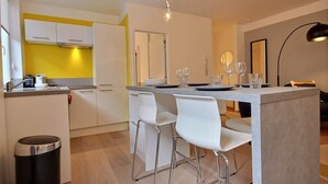 Une cuisine moderne avec des meubles blancs, un mur d’accent jaune et un coin bar doté de chaises et de verres blancs.