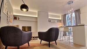Un salon moderne avec deux fauteuils gris, un téléviseur à écran plat et un coin repas équipé d’un bar.