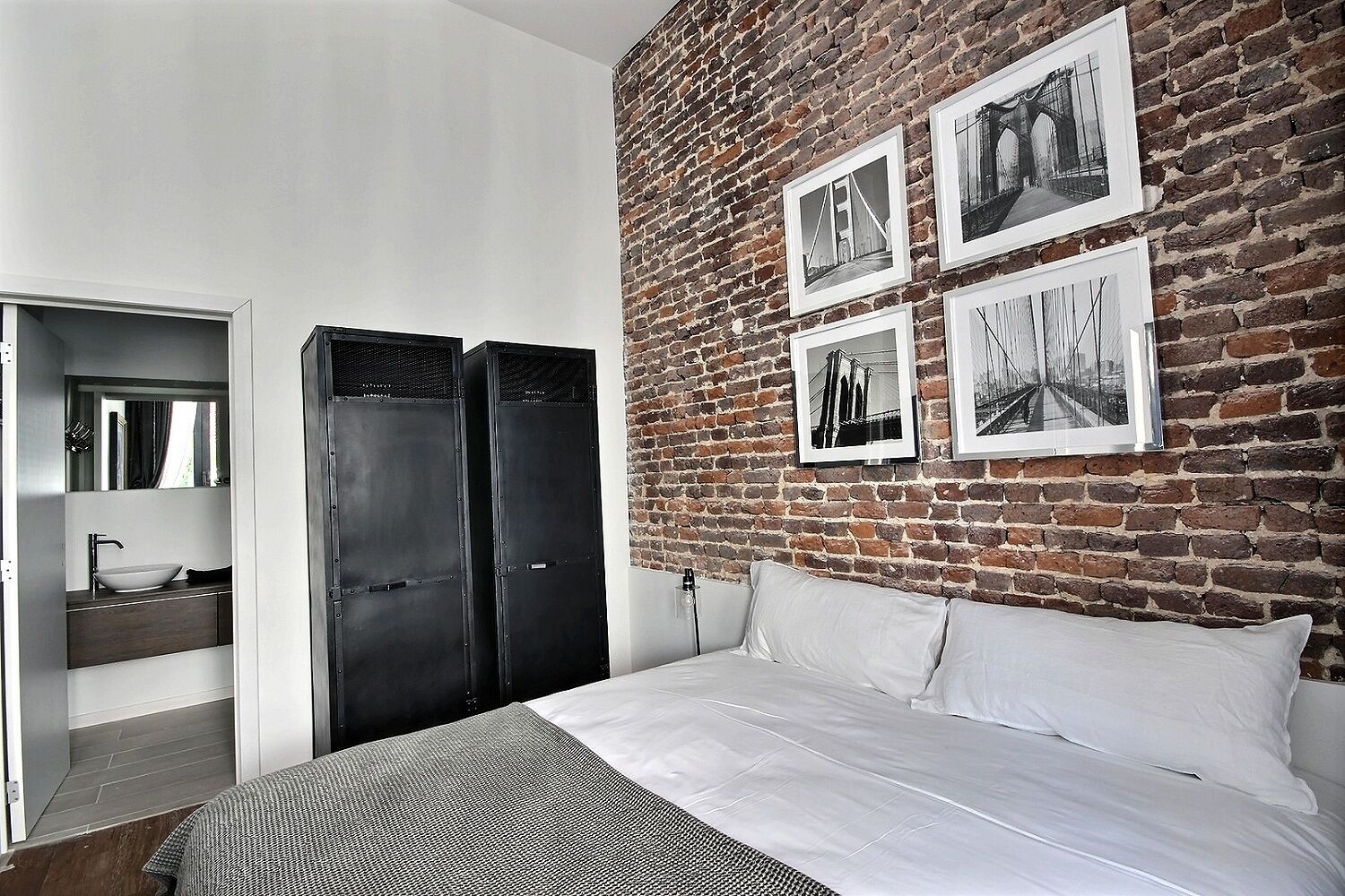 A bedroom with a brick wall, a bed, a nightstand, a mirror, and a wardrobe.