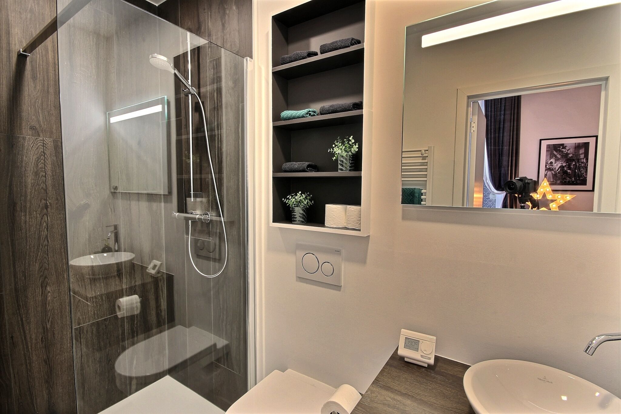 A modern bathroom with a glass-enclosed shower, a vanity with a sink, and a built-in shelf with towels and toiletries.