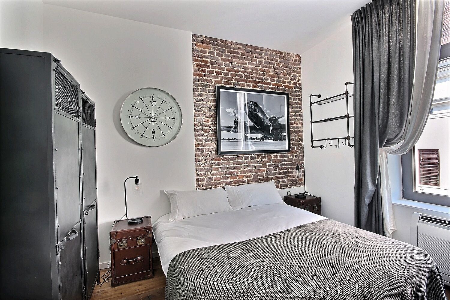 Een slaapkamer met een bakstenen muur, een grote klok, een ingelijst kunstwerk, een bed, een kledingkast en een raam met gordijnen.