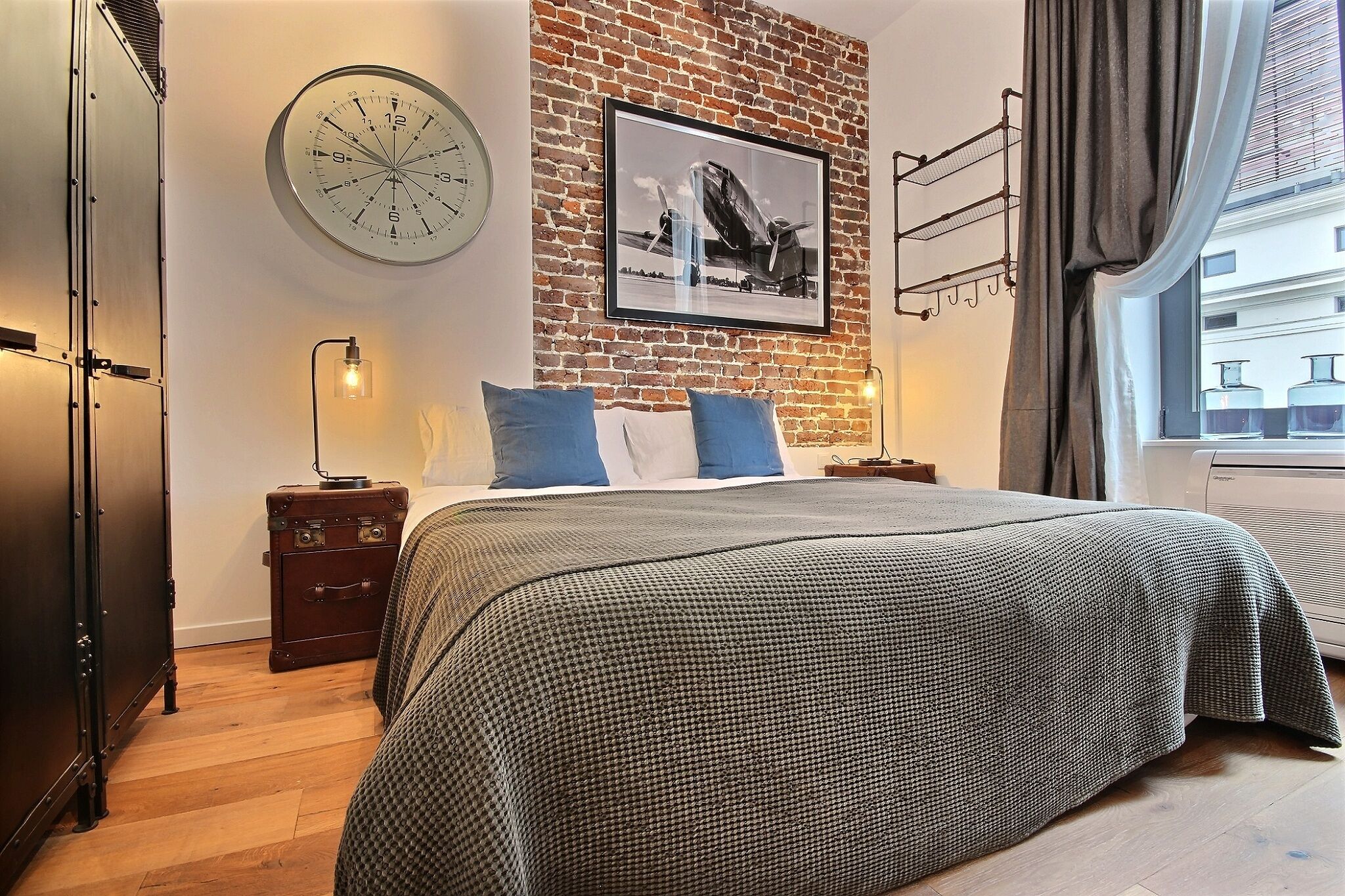 A bedroom with a brick wall, a large clock, a framed picture of an airplane, a bed with a checkered bedspread, and a suitcase on a nightstand.