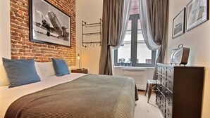 Habitación de hotel con pared de ladrillo, una cama grande con almohadas azules y blancas, un aparador con varios cajones y una ventana con cortinas.