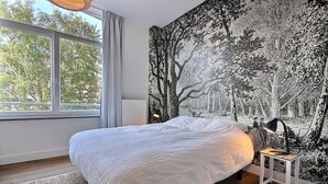 Une chambre avec un grand lit, une fenêtre avec des rideaux, un radiateur et un mur orné d’un motif d’arbre en noir et blanc.