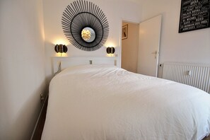 A neatly made bed with a white bedspread, a wall-mounted circular light fixture, and a framed sign listing Parisian landmarks.