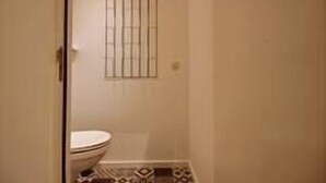 A small bathroom with a toilet, a window with bars, and patterned floor tiles. - Chez Clarisses - 3 bedrooms (Liège)