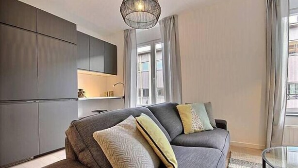 A modern living room with a grey sofa, a glass coffee table, and a kitchen area with grey cabinets. - Chez Clarisses - 3 bedrooms (Liège)