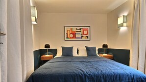 Un lit bien fait, recouvert d’une couette bleue, deux tables de chevet avec des lampes, un tableau abstrait accroché au mur et un rideau de couleur claire.