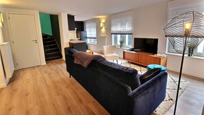 A modern living room with a dark sofa, a white chair, a TV, and a staircase leading to an upper floor. - The KAT105 - 3 bedrooms (Liège)