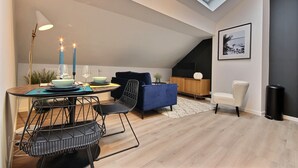 A modern living space with a dining area, a blue sofa, a white chair, and a black trash can. - The KAT106 - 1 bedroom (Liège)